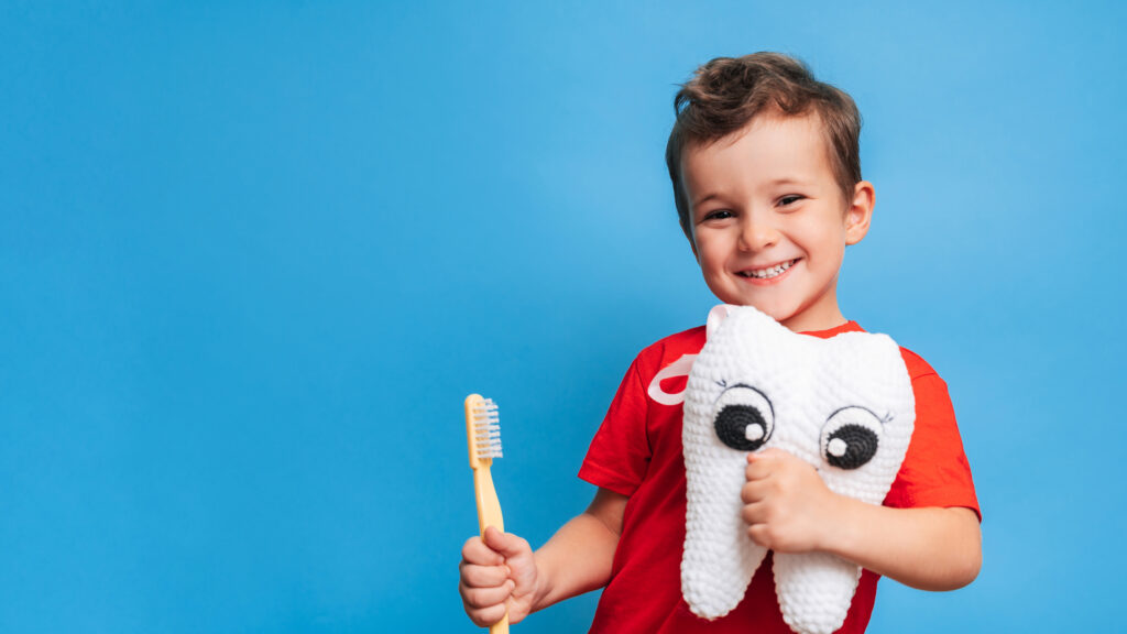 A smiling boy with healthy teeth holds a plush tooth and a toothbrush on a blue isolated background. Oral hygiene. Pediatric dentistry.